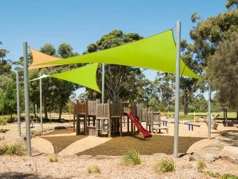 A sunny outdoor playground featuring wooden play equipment under large lime green and tan shade sails