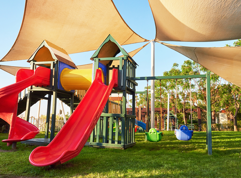 A vibrant playground structure with red slides and colorful swings set against a clear blue sky under sun shades