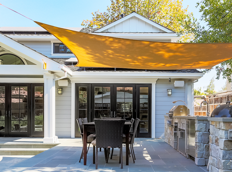 A well-lit backyard patio area featuring a dining set and an outdoor kitchen under a large orange sun shade sail.