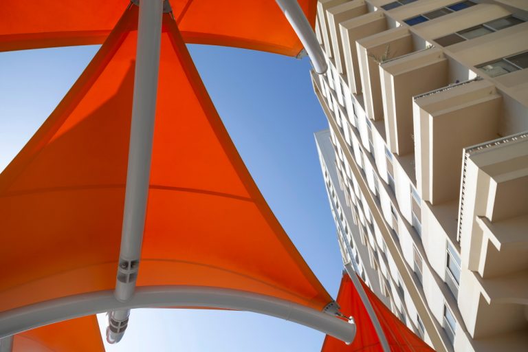 A low-angle upward shot of bright orange tension fabric shade structures against a modern high-rise building